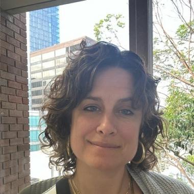 In this selfie, Alex Desautels smiles into the camera. She has short curly brown hair. You can see her shoulders -- she is wearing a black and white top and gray cardigan. She is in front of a window and tree branches and a brick building are visible in the background.
